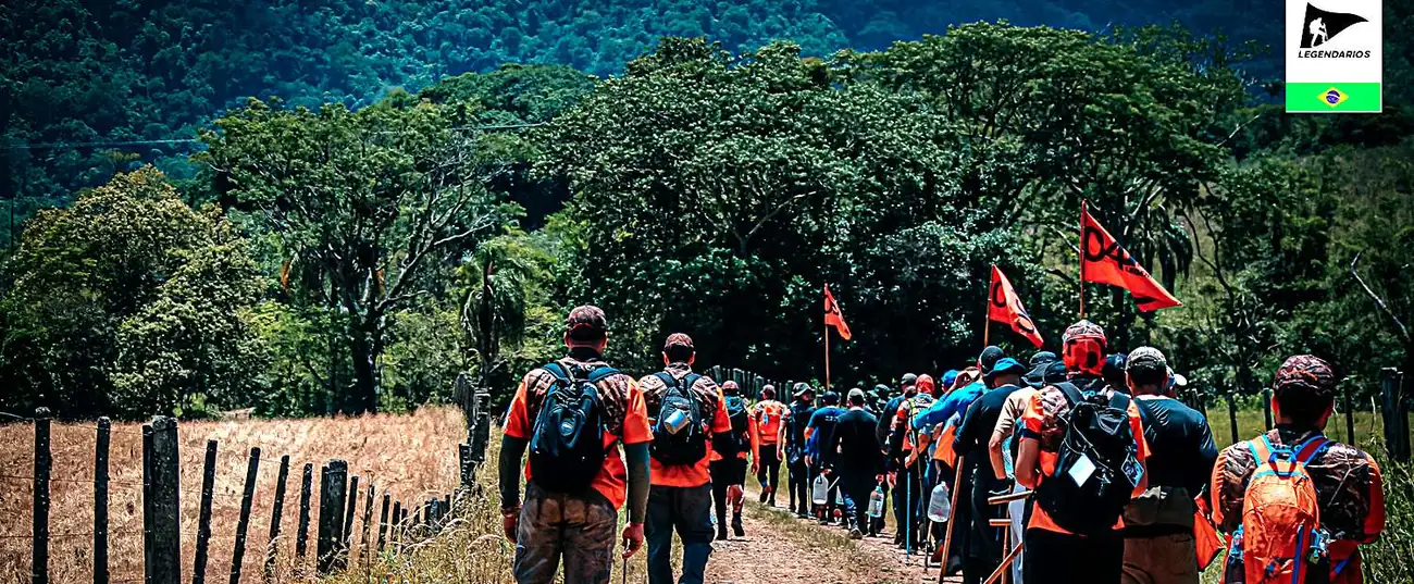 Fila de homens caminhando numa estrada de chão, com as bandeiras do Legendários